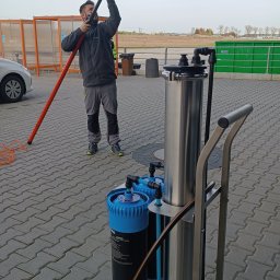 Clean Case Sp. z o.o. - Mężczyzna w szarym ubraniu obsługuje urządzenie do mycia okien na wysięgniku z systemem filtracji wody, widoczne filtry i metalowa konstrukcja wózka, na zewnątrz, kostka brukowa.