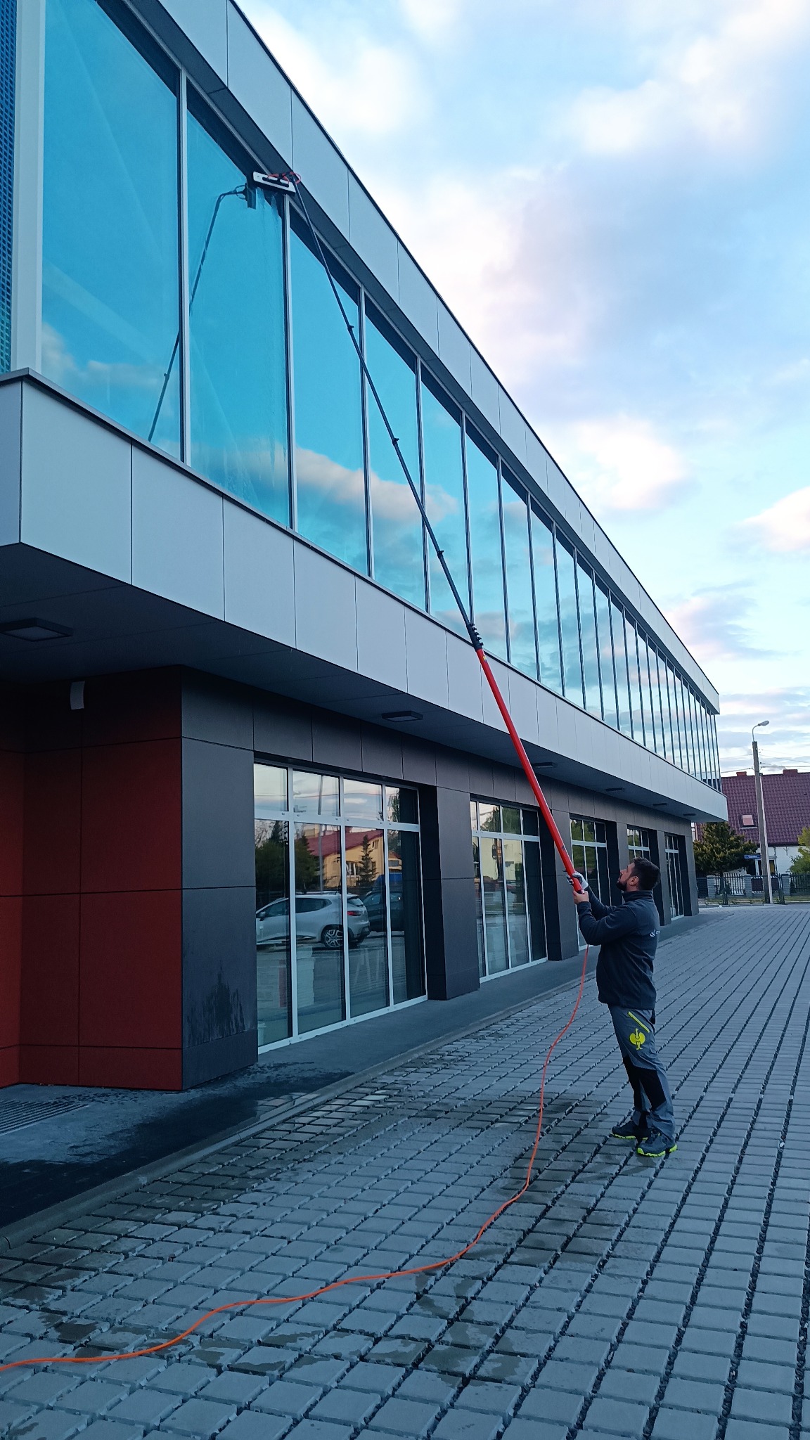 Pracownik w szarym kombinezonie myje okna biurowca za pomocą długiej, czerwonej lancy z zamontowaną szczotką; na chodniku widoczny pomarańczowy wąż doprowadzający wodę.