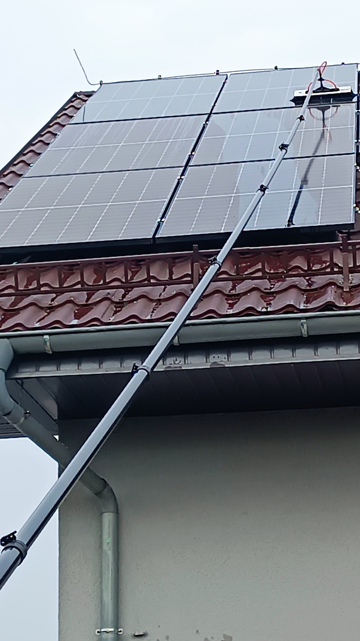 Czyszczenie paneli słonecznych na dachu z użyciem teleskopowej szczotki zraszającej, widoczne brązowe dachówki i szara elewacja budynku.