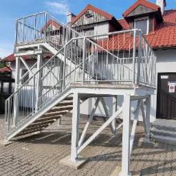 Zewnętrzne, metalowe schody przeciwpożarowe z platformą, prowadzące do budynku w Głodowie, z widocznymi znakami bezpieczeństwa na drzwiach.