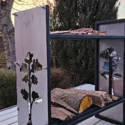 Metalowy regał na drewno z wyciętym laserowo motywem drzewa, stojący na blaszanym dachu, z widocznym drewnem opałowym na półkach, na tle drzew i zachodzącego słońca.