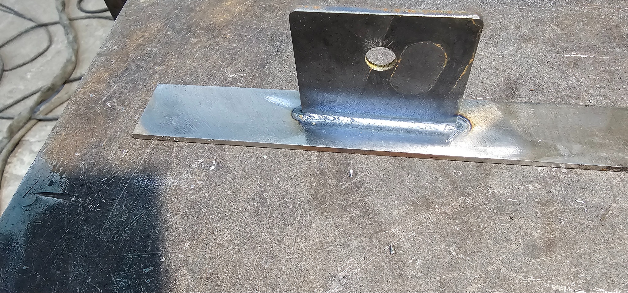 Spoina łącząca płaskownik z pionowym elementem z otworem, wykonana na szorstkim metalowym stole.