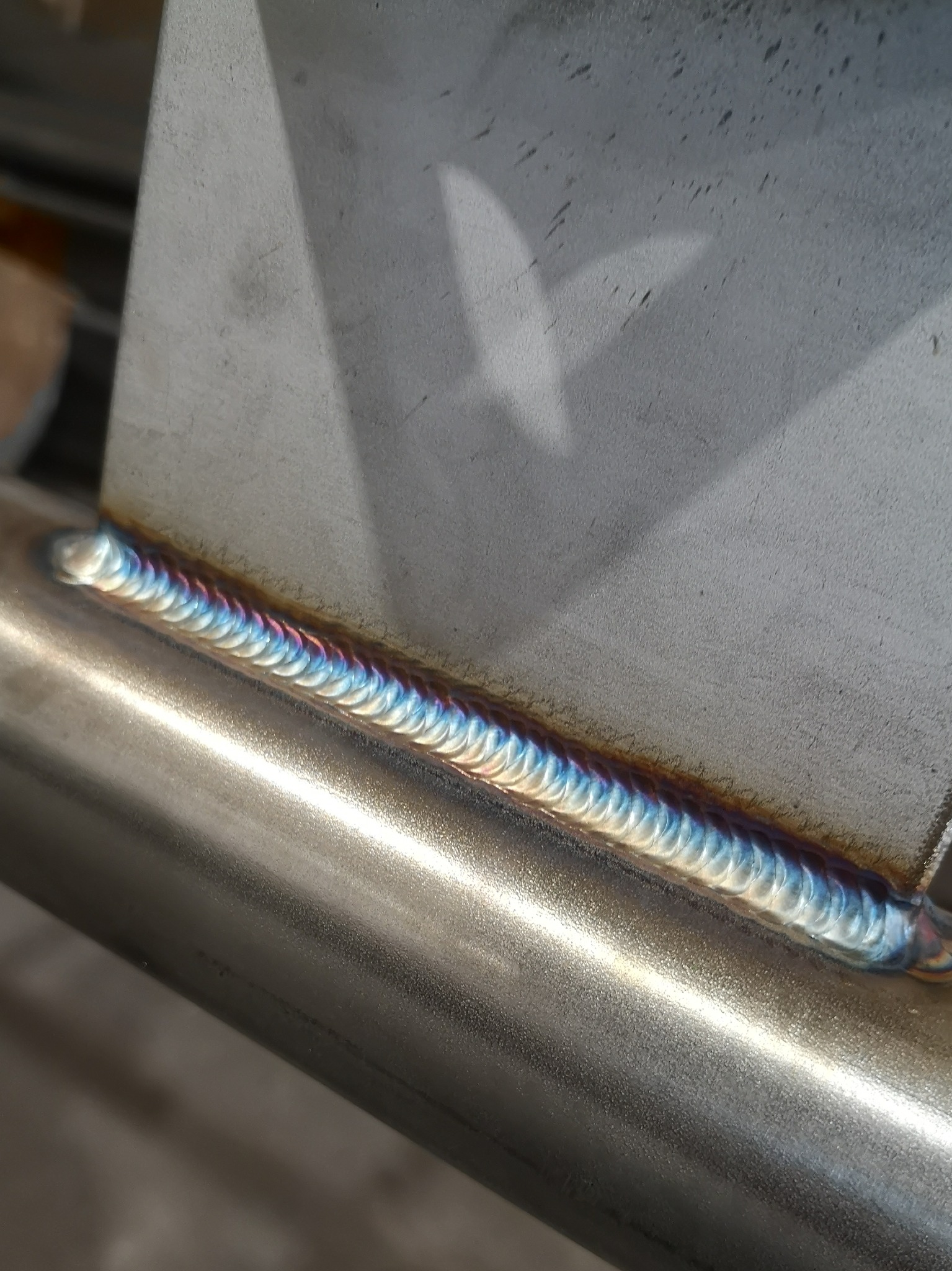 Metalowa konstrukcja po spawaniu, widoczny precyzyjny szew spawalniczy z charakterystycznymi kolorami utlenionego metalu, łączący rurę z płaskownikiem.