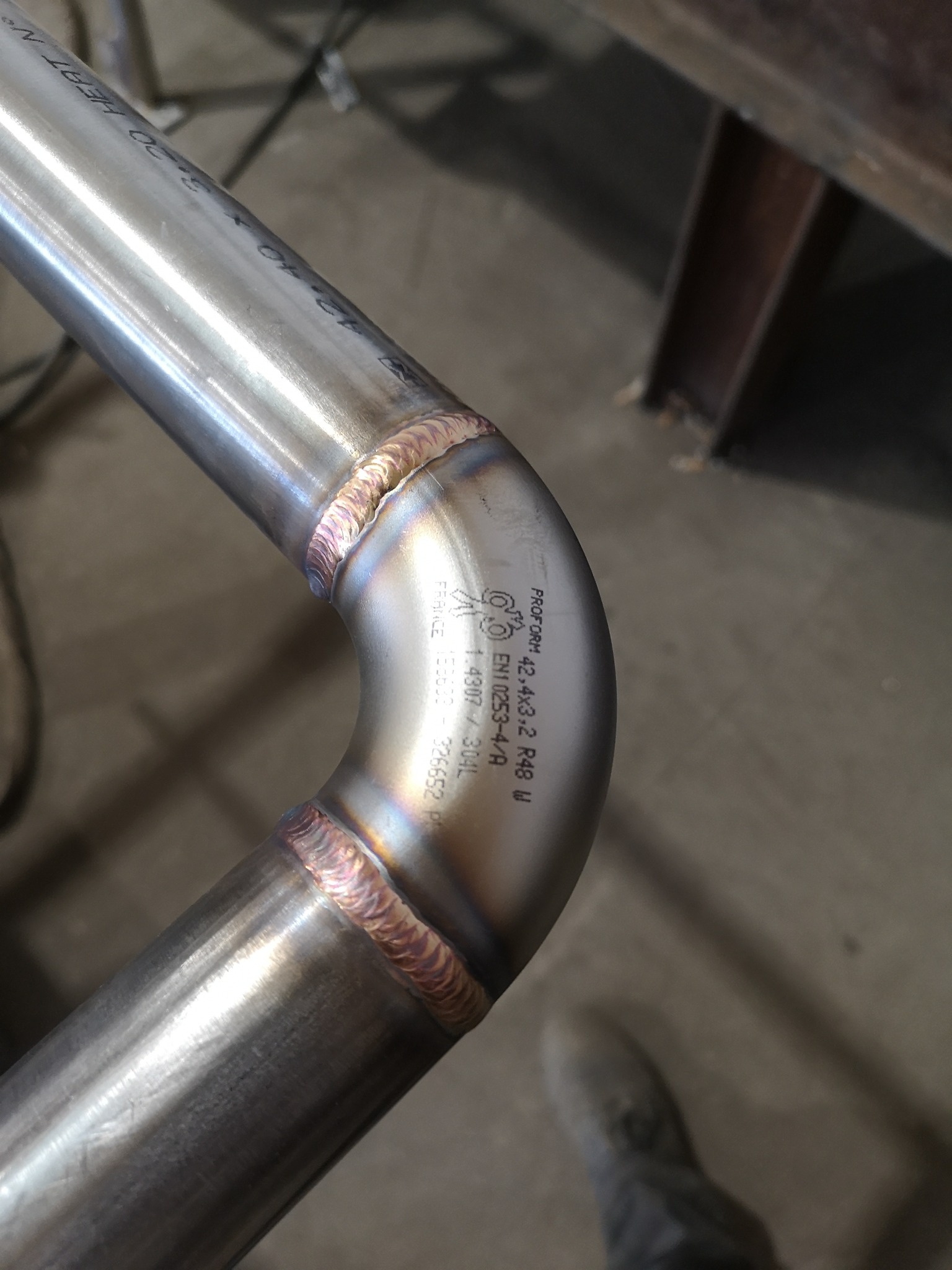 Precyzyjne spawy na rurze ze stali nierdzewnej, widoczne charakterystyczne ślady po spawaniu TIG, z oznaczeniami producenta na powierzchni rury.