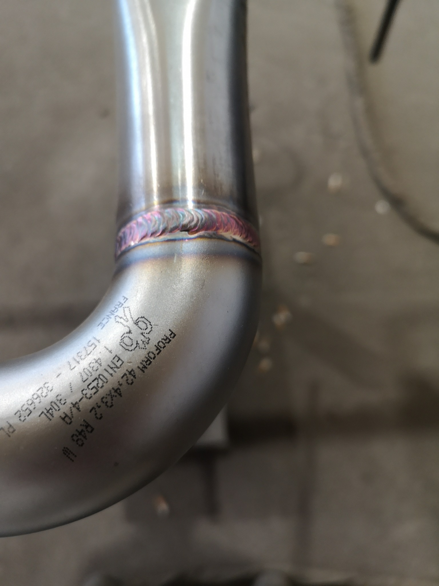 Szczegółowe zbliżenie na spawany złącze rury ze stali nierdzewnej, widoczne ślady spawania TIG i laserowy nadruk z oznaczeniami technicznymi.