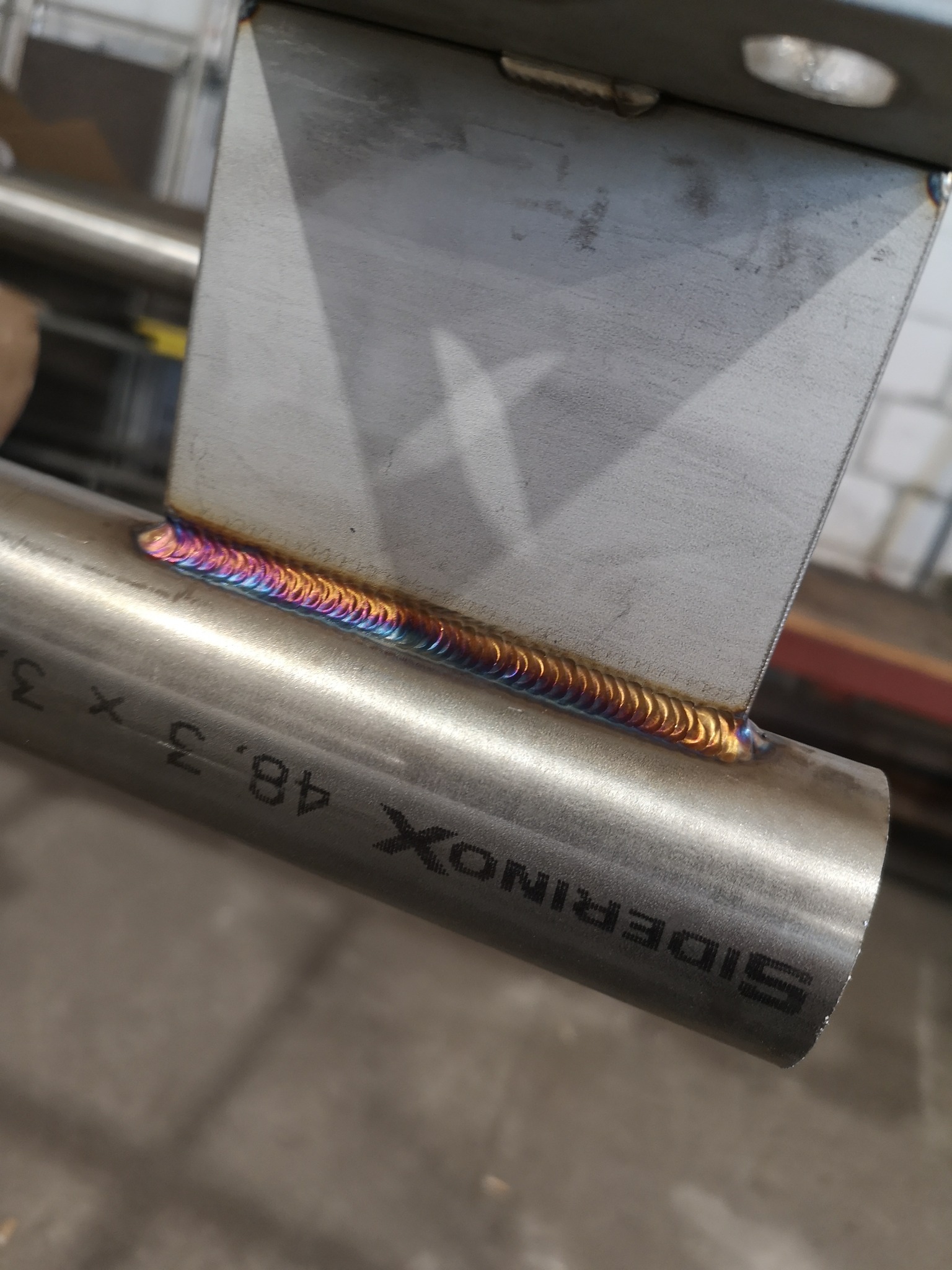 Szlifowana rura ze stali nierdzewnej z przyspawanym elementem, widoczna tęczowa spoina TIG z bliska i napis SIDERINOX 48.3 x 3