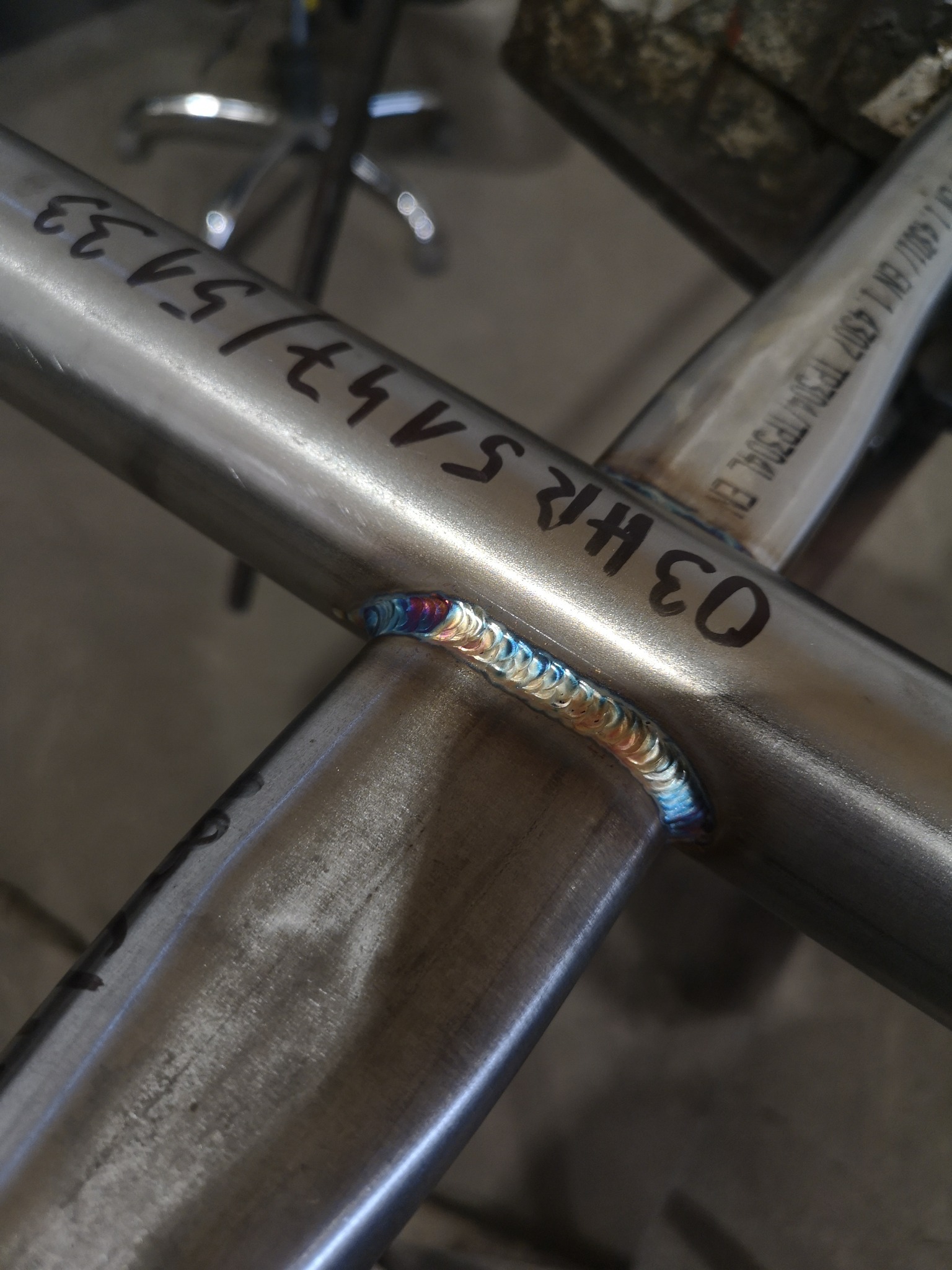 Spoina spawalnicza łącząca dwie rury ze stali nierdzewnej, widoczne charakterystyczne kolory przegrzanego metalu, na rurach naniesione czarnym markerem oznaczenia.