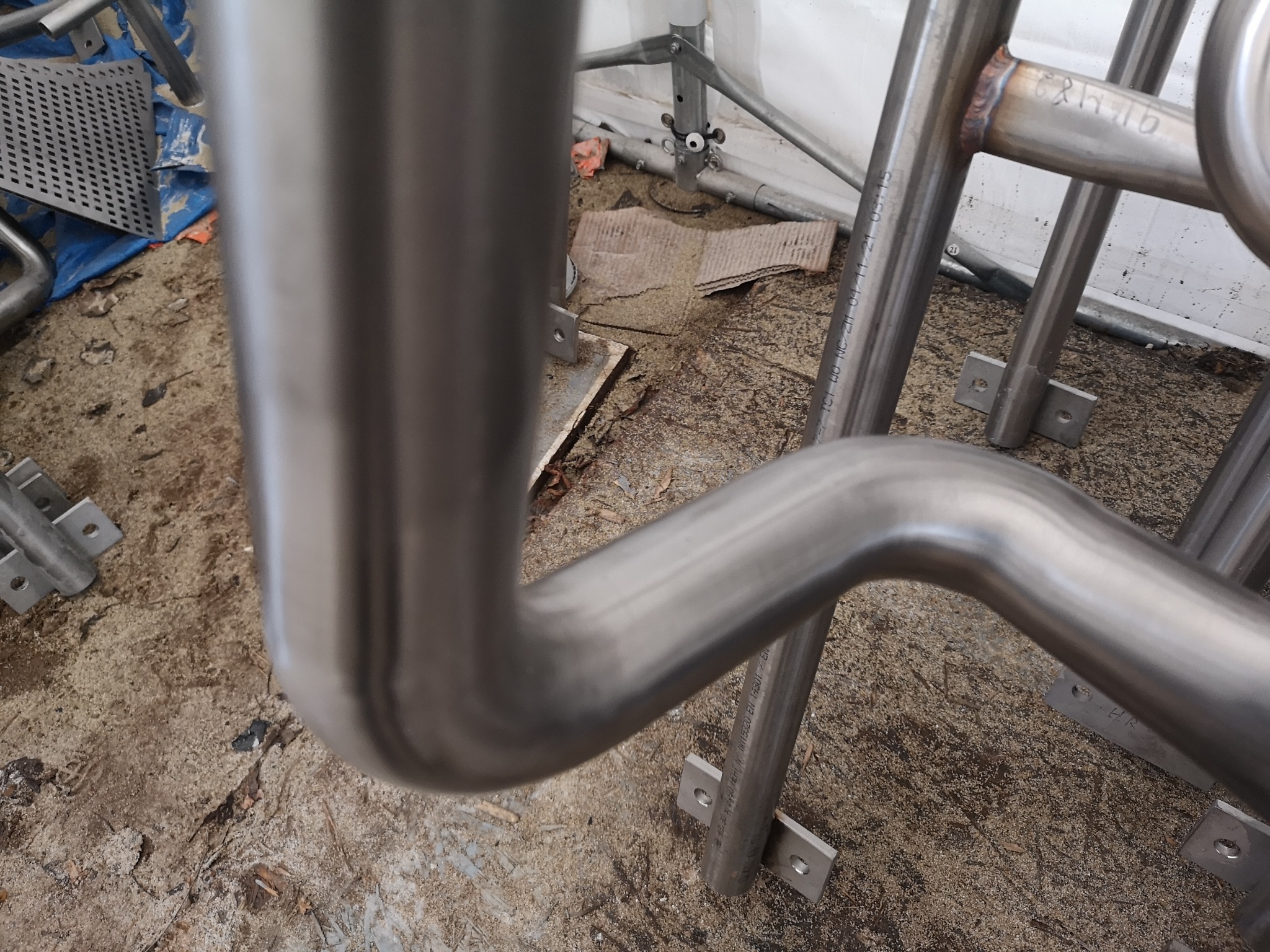 Metalowa konstrukcja ze spawanych rur, z widocznymi spawami i oznaczeniami producenta, w surowym otoczeniu warsztatowym.