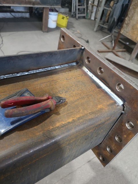 Detale świeżo pospawanej, pomalowanej podkładem konstrukcji stalowej w warsztacie, z widocznymi spawami i śladami korozji, obok leżą kombinerki.