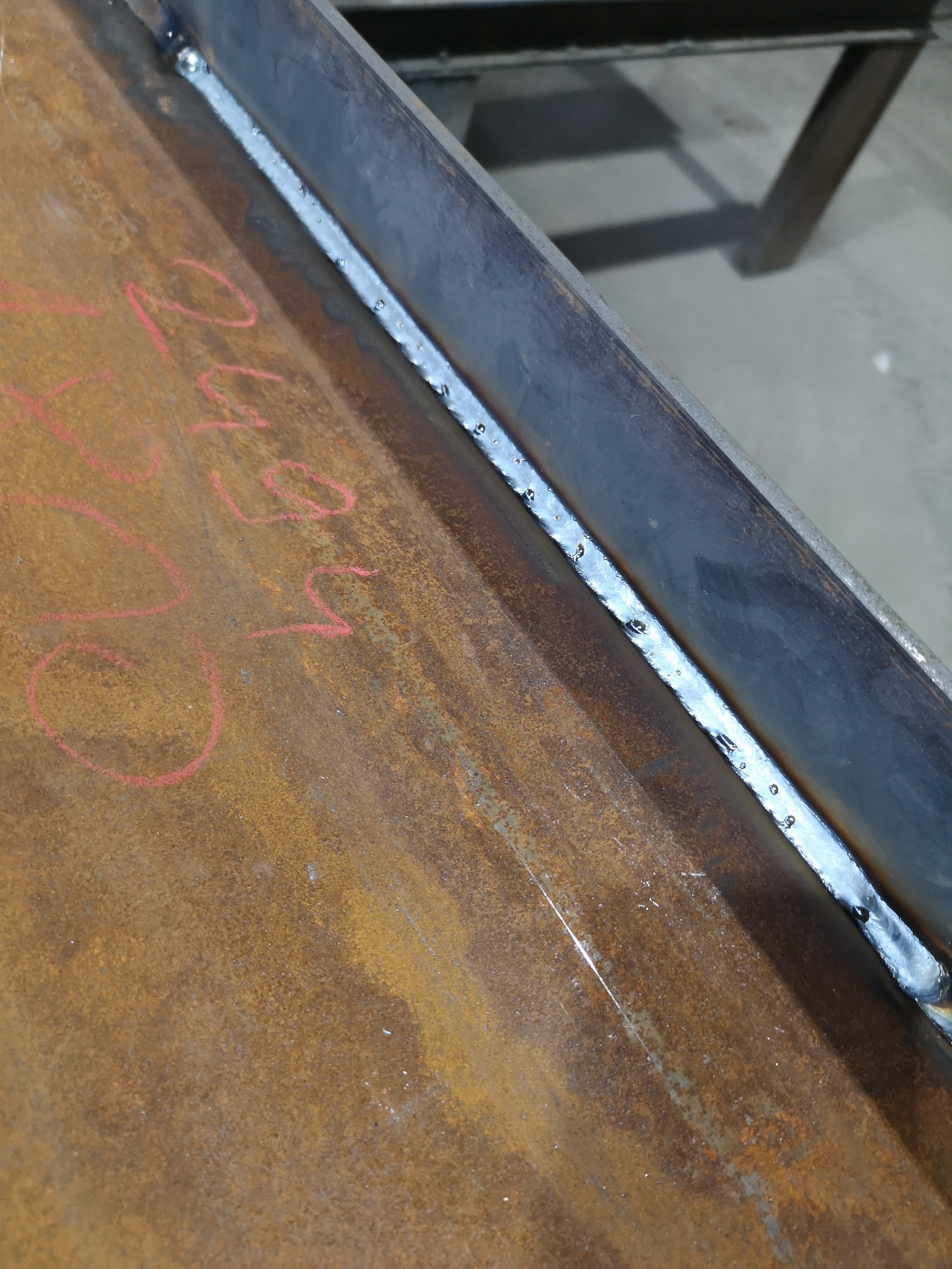 Świeżo wykonany spaw na zardzewiałej, metalowej belce z widocznymi śladami po spawaniu i napisem wykonanym czerwoną kredą.