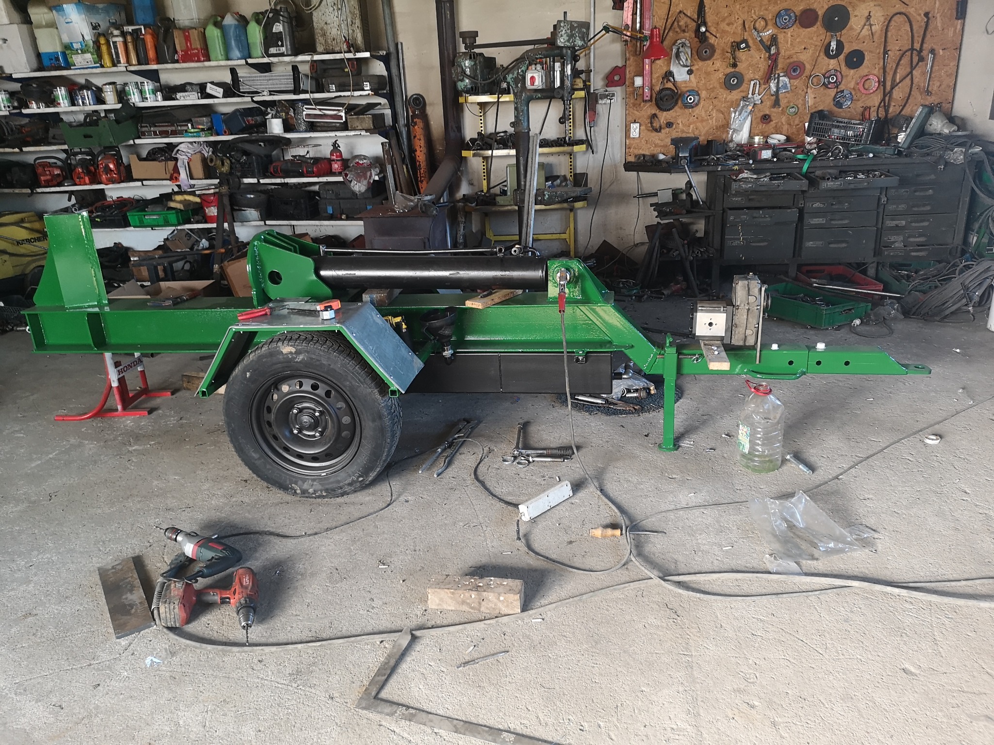 Zielony, metalowy rozłupywacz do drewna na przyczepce, w warsztacie z narzędziami i półkami pełnymi pojemników, w trakcie budowy, widoczne wiertarki i elementy metalowe na posadzce.