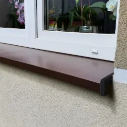 Brązowy parapet zewnętrzny zamontowany pod białym oknem na elewacji budynku o jasnej, ziarnistej strukturze.