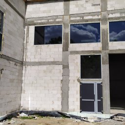 OKNOMAG Krzysztof Niemyjski - Surowy budynek w trakcie budowy z zamontowanymi oknami i drzwiami, widoczne pianki montażowe, odbicie chmur w szybach.