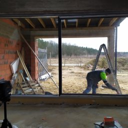 OKNOMAG Krzysztof Niemyjski - Montaż przesuwnych okien tarasowych w surowym stanie, widoczny pracownik na drabinie i laserowy niwelator na statywie.