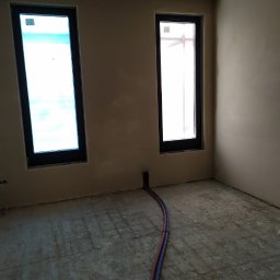 OKNOMAG Krzysztof Niemyjski - Surowy stan wnętrza z dwoma pionowymi oknami w ciemnych ramach, widoczne wystające rury instalacyjne z podłogi.