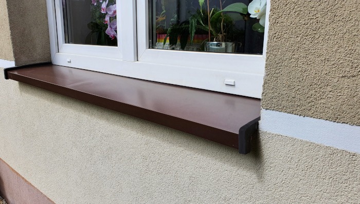 Brązowy parapet zewnętrzny zamontowany pod białym oknem na elewacji budynku o jasnej, ziarnistej strukturze.