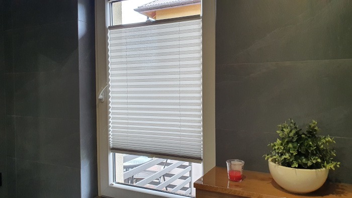 Jasne plisy okienne zamontowane w oknie, widok na balkon z metalową barierką, obok dekoracja roślinna w białej doniczce i świeczka w szklanym świeczniku, ciemna ściana w tle.