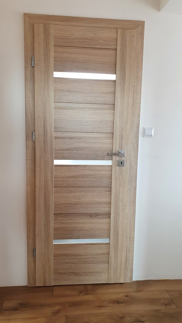 Zamknięte drzwi wewnętrzne z drewnopodobną okleiną, z poziomymi przeszkleniami, zamontowane w białej ścianie, widoczny fragment podłogi.
