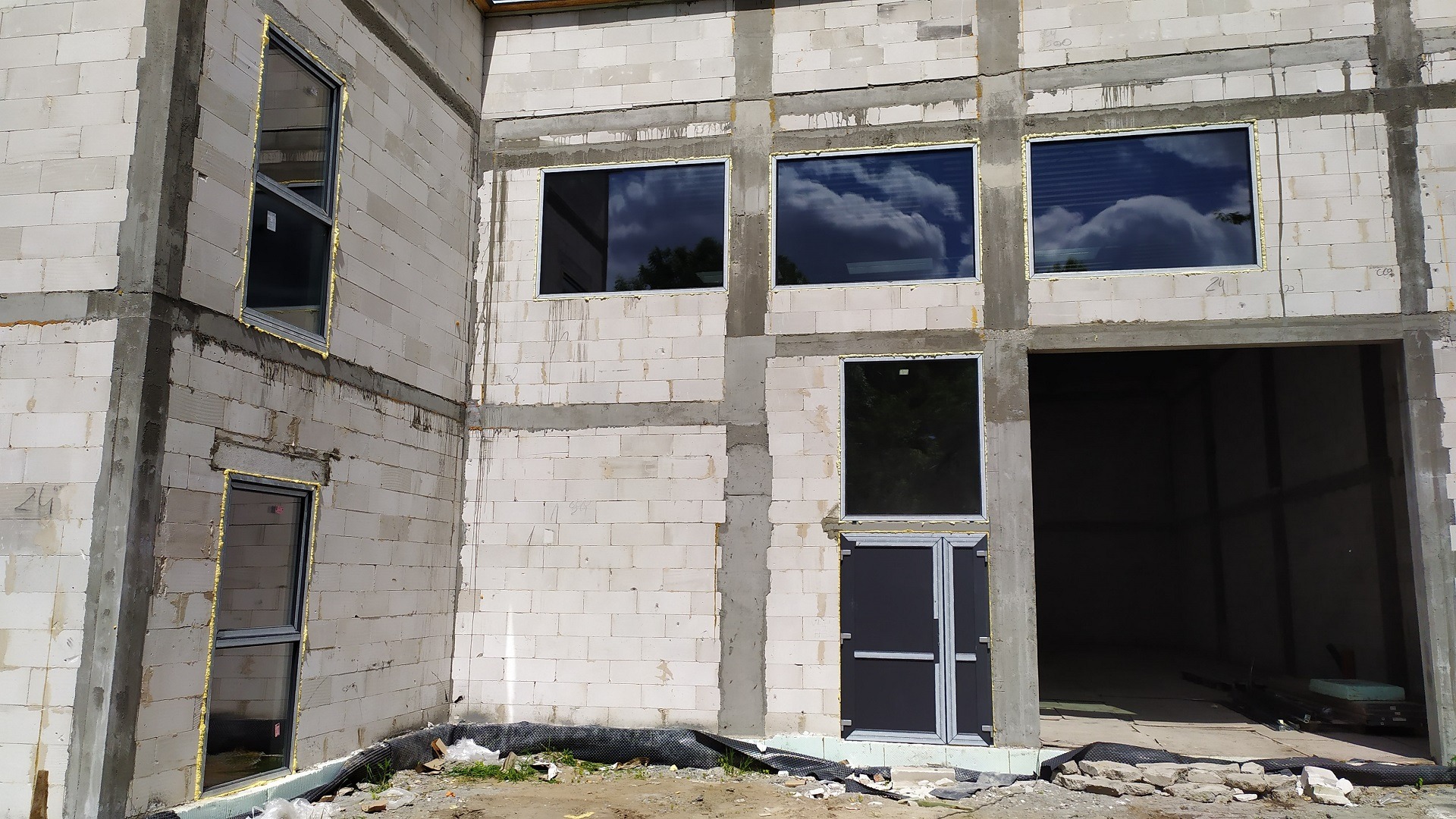 Surowy budynek w trakcie budowy z zamontowanymi oknami i drzwiami, widoczne pianki montażowe, odbicie chmur w szybach.