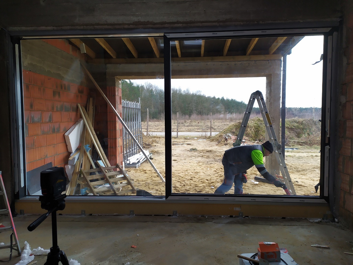 Montaż przesuwnych okien tarasowych w surowym stanie, widoczny pracownik na drabinie i laserowy niwelator na statywie.