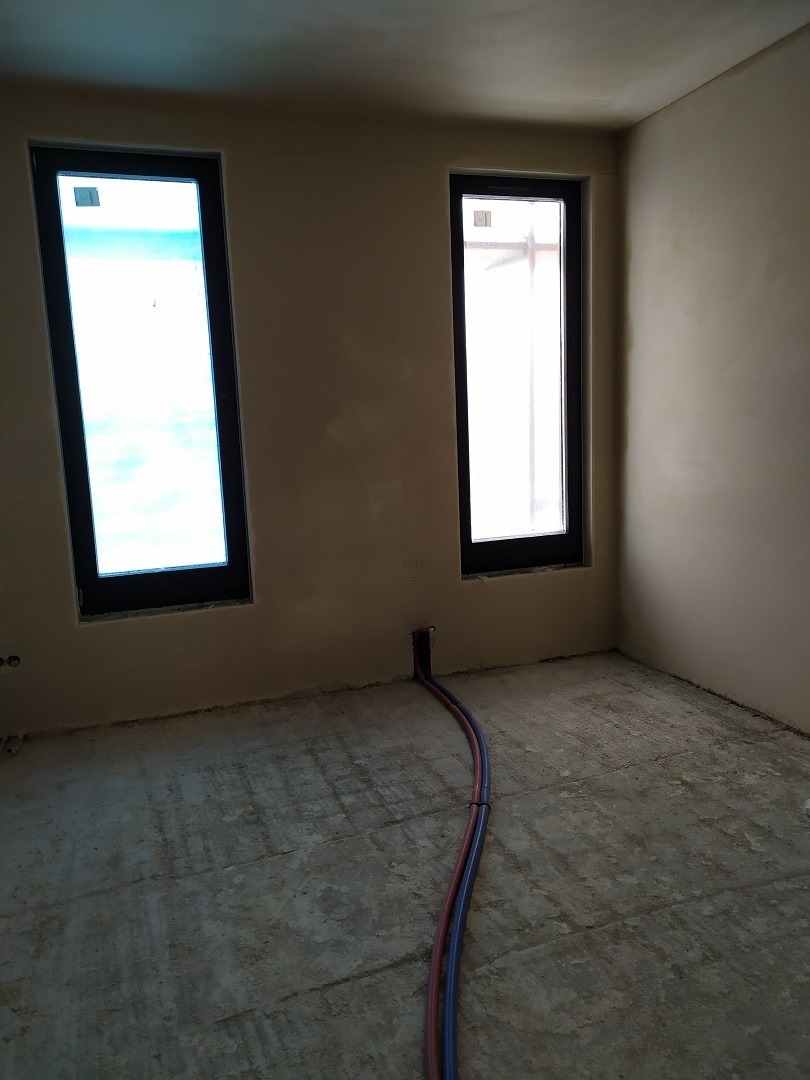 Surowy stan wnętrza z dwoma pionowymi oknami w ciemnych ramach, widoczne wystające rury instalacyjne z podłogi.