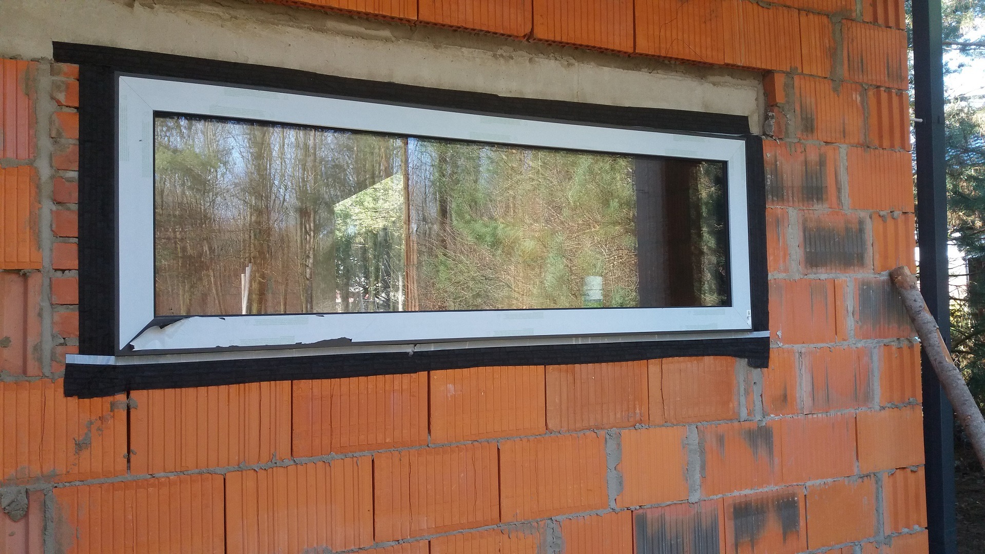Nowe białe okno PCV osadzone w surowej, pomarańczowej ścianie z pustaków, z widoczną czarną taśmą uszczelniającą i folią ochronną na ramie, odzwierciedlające drzewa w szybie.