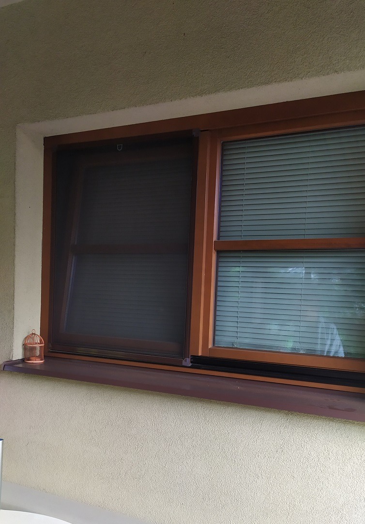 Brązowe okno z przesuwaną moskitierą i żaluzjami poziomymi, na parapecie dekoracyjna klatka dla ptaków