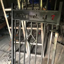 Metalowa furtka z napisem 'Piotrówek II 7' w warsztacie, widoczne elementy konstrukcyjne i narzędzia w tle.