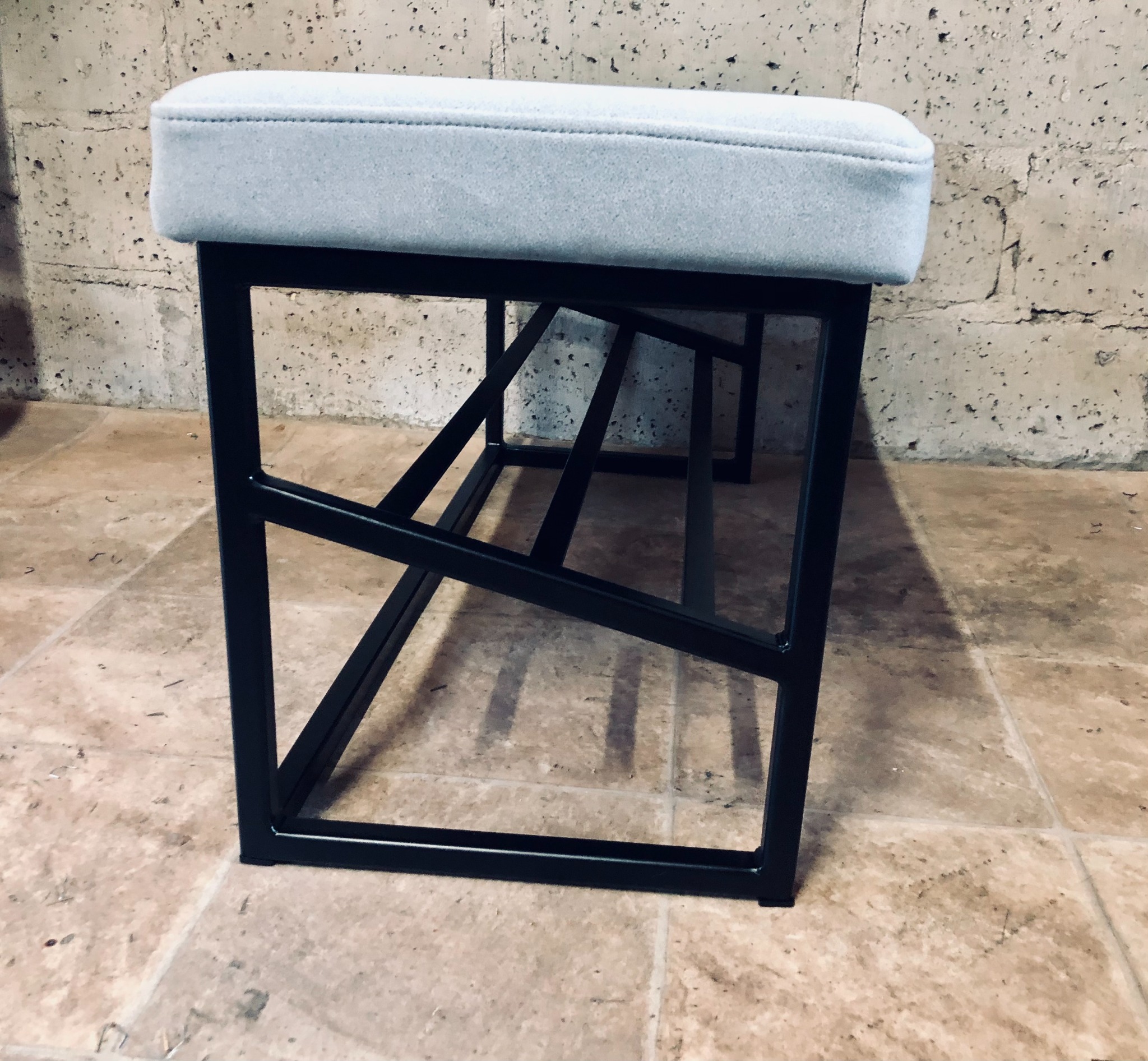 Nowoczesny taboret z czarną metalową ramą i jasnoszarym, tapicerowanym siedziskiem, ustawiony na kafelkach w surowym wnętrzu z betonową ścianą w tle.