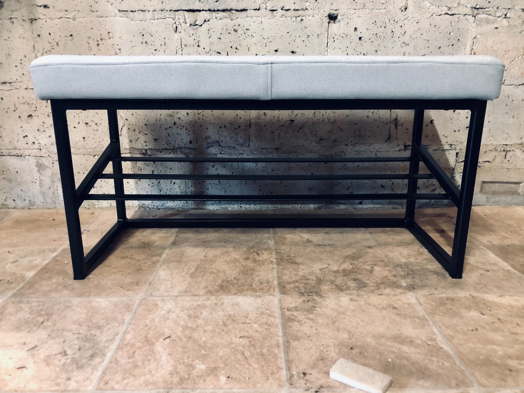 Metalowa ławka z szarą tapicerką siedziska i półkami na buty, ustawiona na kafelkach przed surową, betonową ścianą.