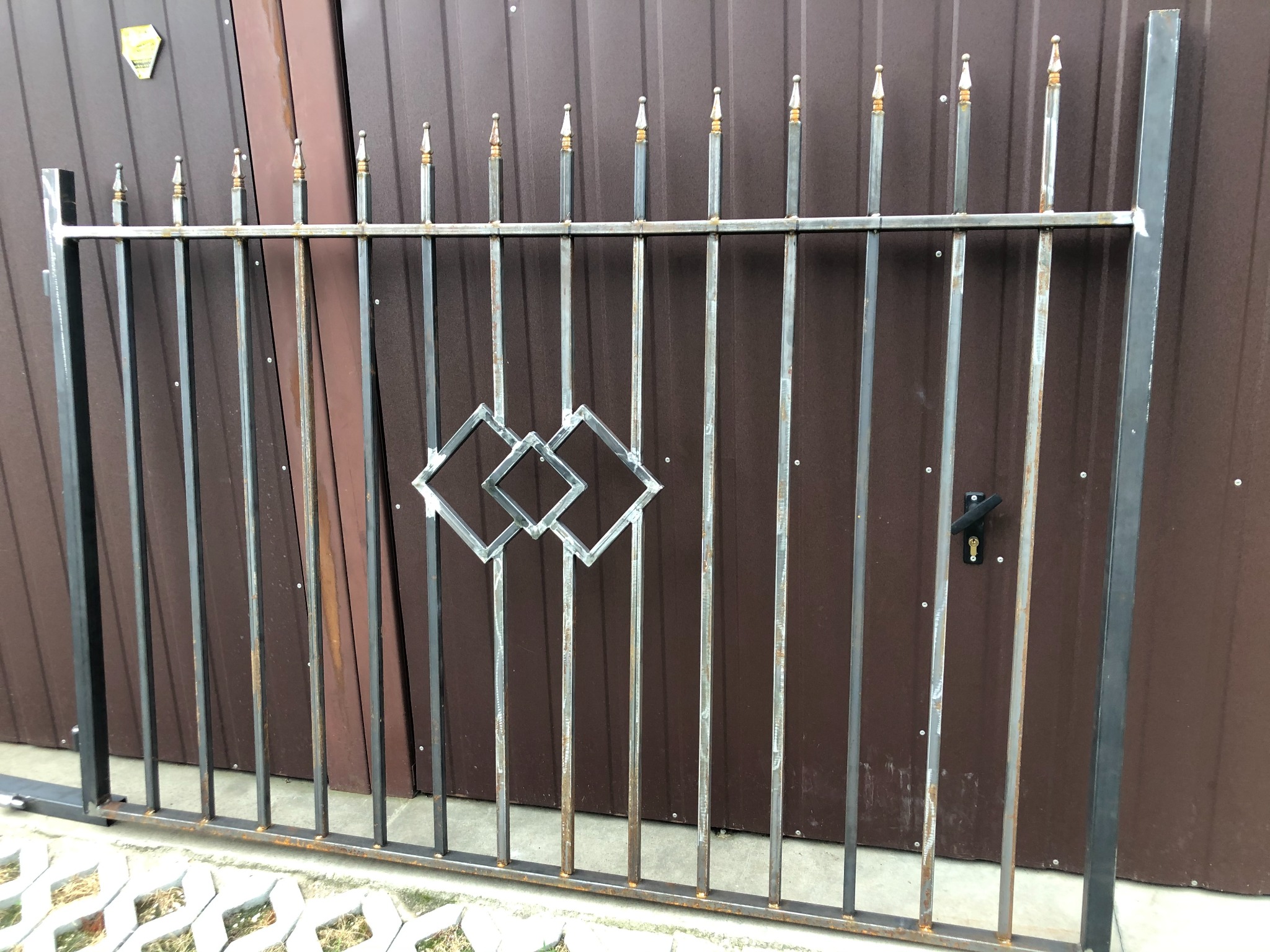 Surowa, metalowa konstrukcja bramy wjazdowej z ozdobnymi grotami i motywem trzech rombów, oparta o brązową blachę falistą.