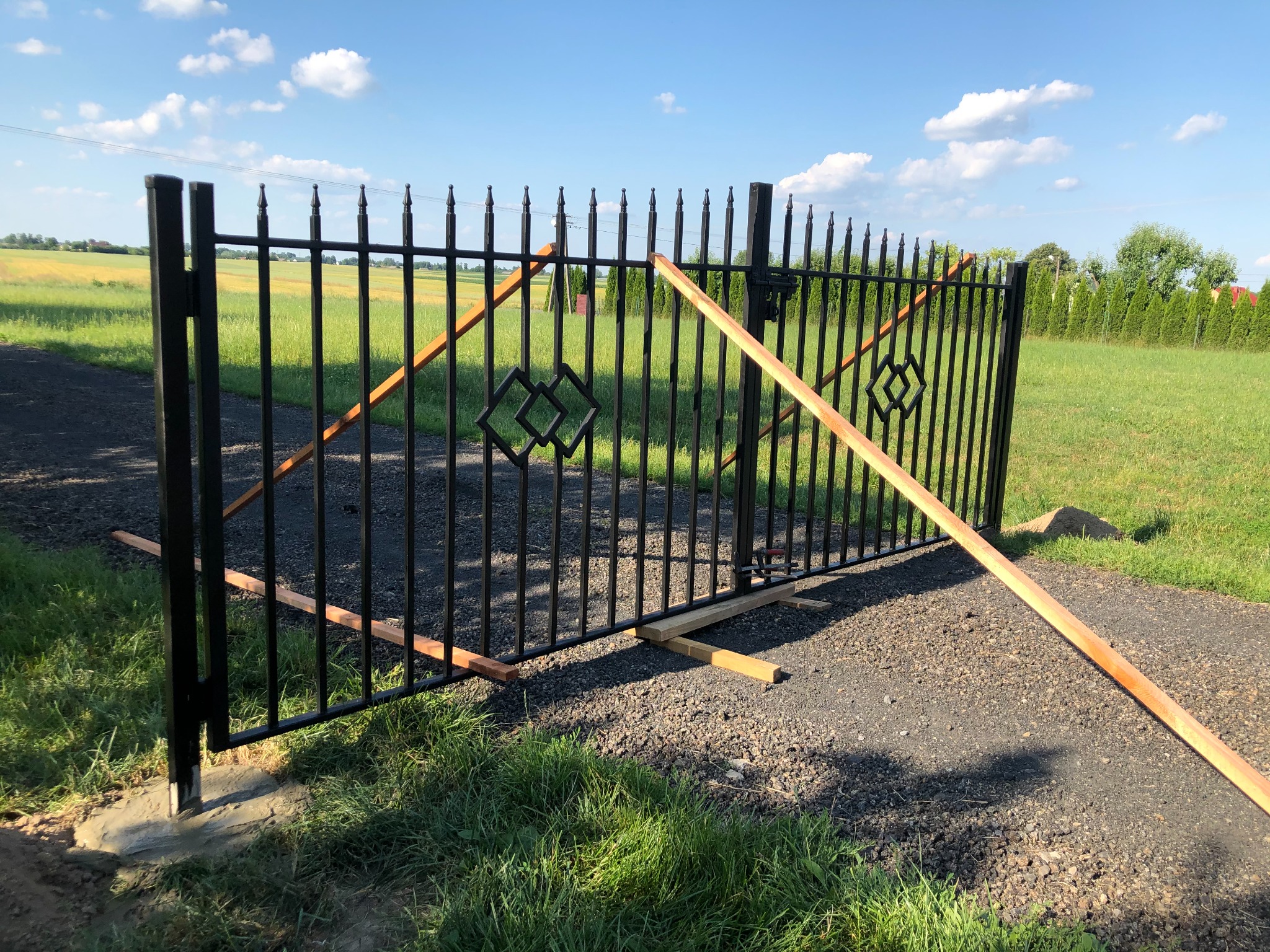 Czarna metalowa brama wjazdowa z dekoracyjnymi elementami w trakcie montażu, podparta drewnianymi listwami, w tle zielony trawnik i pole.