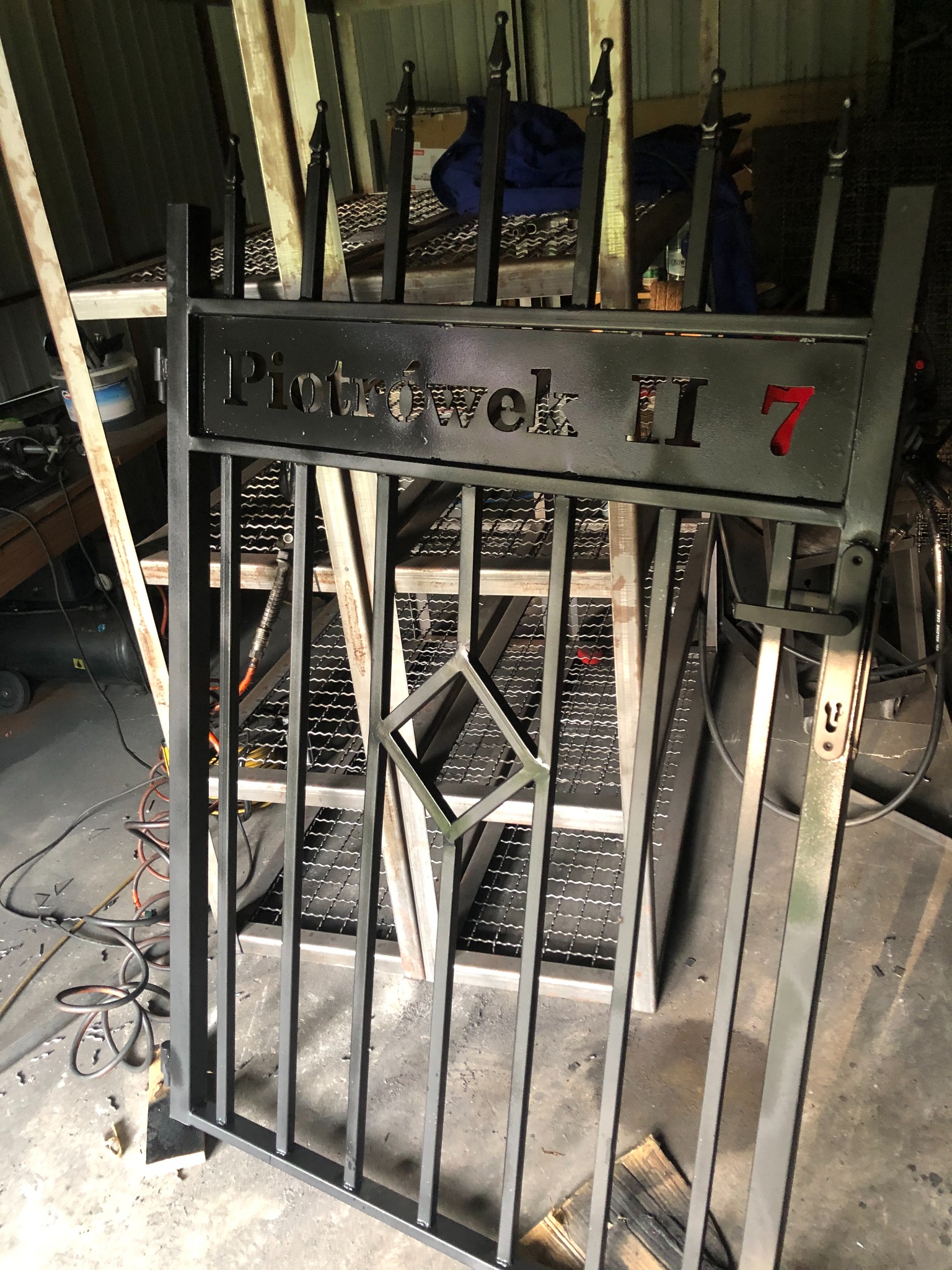Metalowa furtka z napisem 'Piotrówek II 7' w warsztacie, widoczne elementy konstrukcyjne i narzędzia w tle.