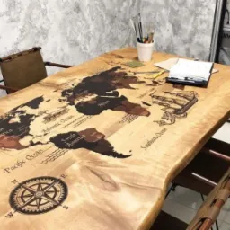 Drewniany stół z nieregularnymi krawędziami, zdobiony mapą świata i kompasem, otoczony krzesłami z płóciennymi oparciami.