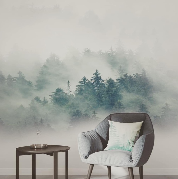 Pokój z fototapetą przedstawiającą mglisty las iglasty, szary fotel z poduszką z motywem lasu, stolik kawowy z dekoracją.