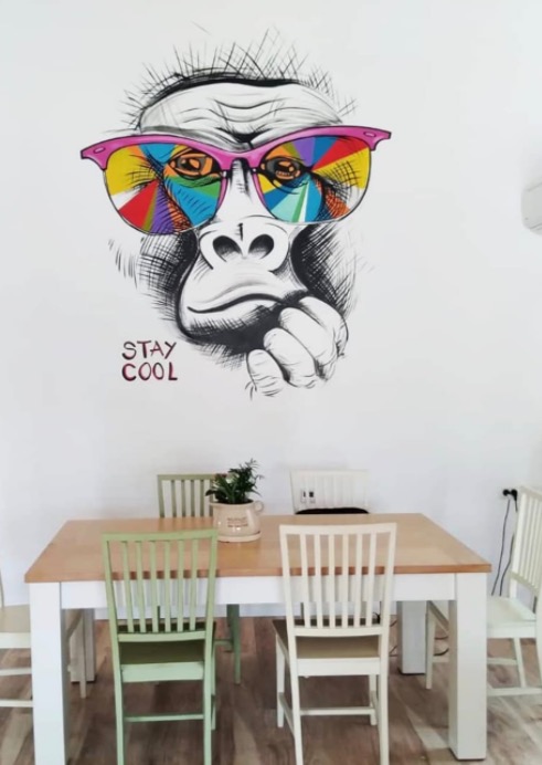 Ściana w jadalni ozdobiona czarno-białym rysunkiem goryla w różowych okularach z kolorowymi szkłami, pod rysunkiem napis 'STAY COOL', poniżej stół z czterema krzesłami.