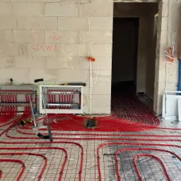 Rozłożone czerwone rury ogrzewania podłogowego na siatce, widoczne rozdzielacze w skrzynkach, ściany w surowym stanie z napisami, narzędzia pracy w tle.