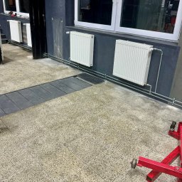 ARISTOCLAN STUDIO SPÓŁKA Z OGRANICZONĄ ODPOWIEDZIALNOŚCIĄ - Białe grzejniki panelowe zamontowane na szarej ścianie, z widocznymi rurami i zaworami, na podłodze z terakoty. W tle okno i czarny portal drzwiowy. W rogu czerwony stojak warsztatowy na kółkach.