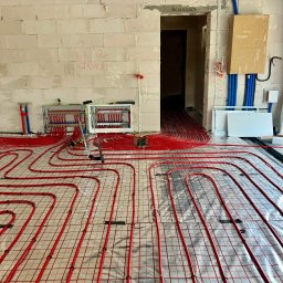 ARISTOCLAN STUDIO SPÓŁKA Z OGRANICZONĄ ODPOWIEDZIALNOŚCIĄ - Rozłożone czerwone rury ogrzewania podłogowego na srebrnej folii z siatką, widoczne rozdzielacze i instalacje na ścianie z białych bloczków w surowym stanie.