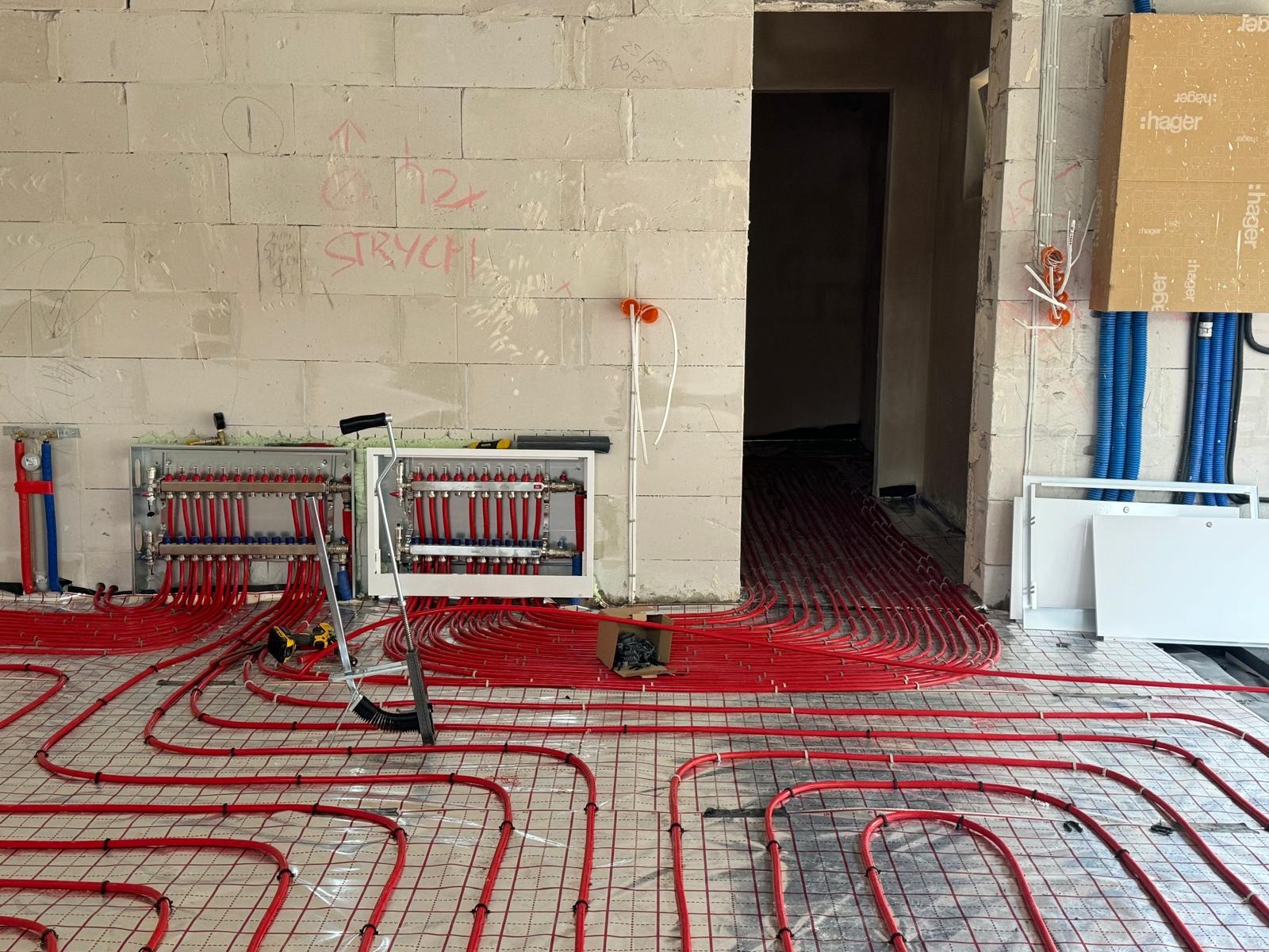 Rozłożone czerwone rury ogrzewania podłogowego na siatce, widoczne rozdzielacze w skrzynkach, ściany w surowym stanie z napisami, narzędzia pracy w tle.