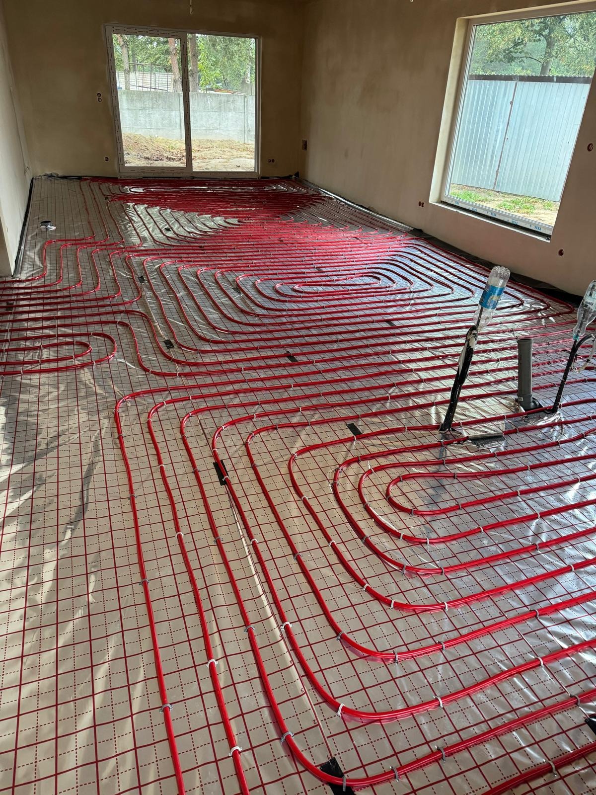Wnętrze w stanie surowym z rozłożoną instalacją ogrzewania podłogowego z czerwonych rur na siatce, widoczne okna i butelki z wodą.