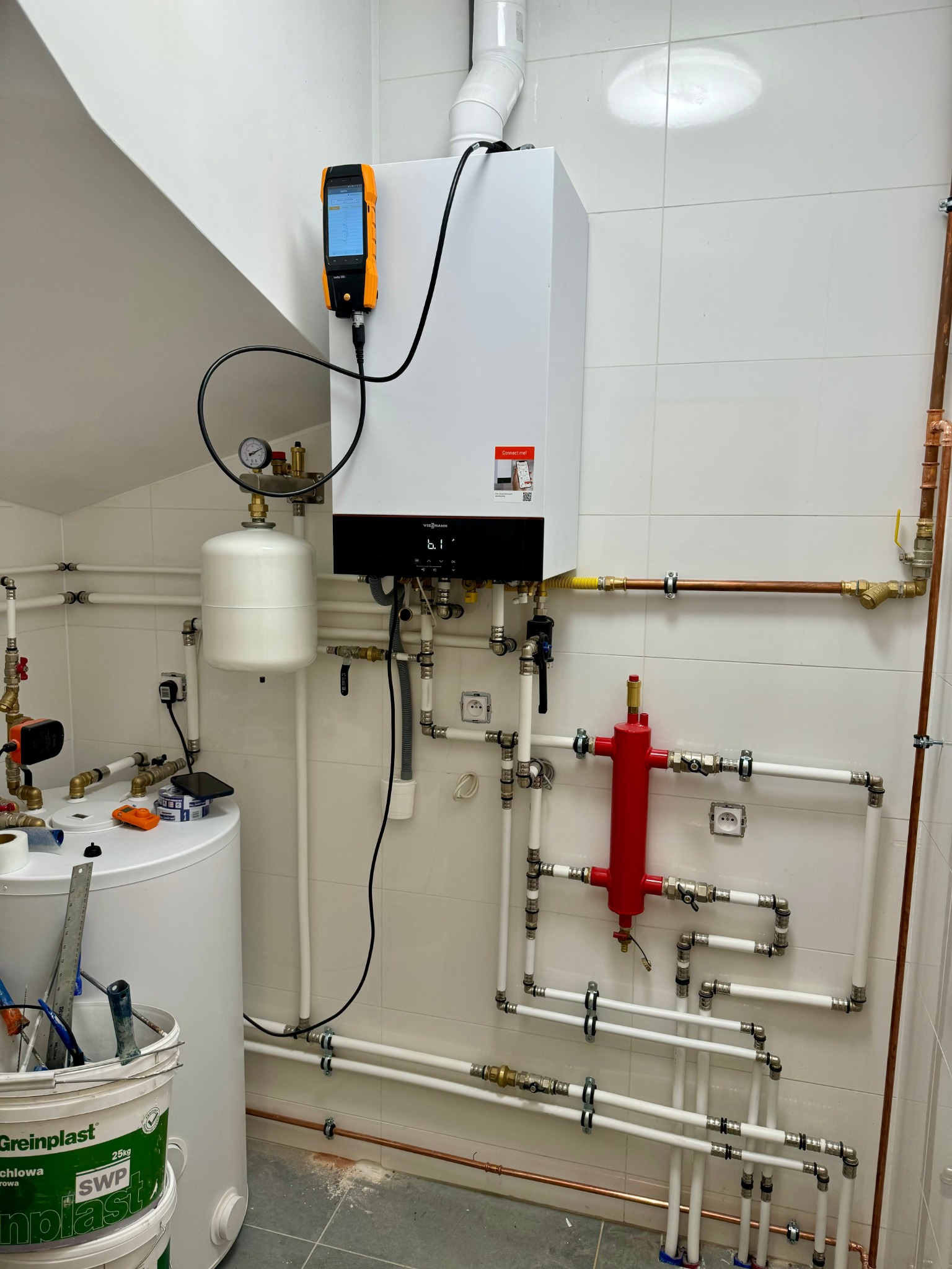 Biały piec grzewczy Viessmann z podłączonym analizatorem spalin, widoczne rury miedziane i plastikowe, oraz biały zbiornik wyrównawczy. Na podłodze wiadro z farbą Greinplast i narzędzia.