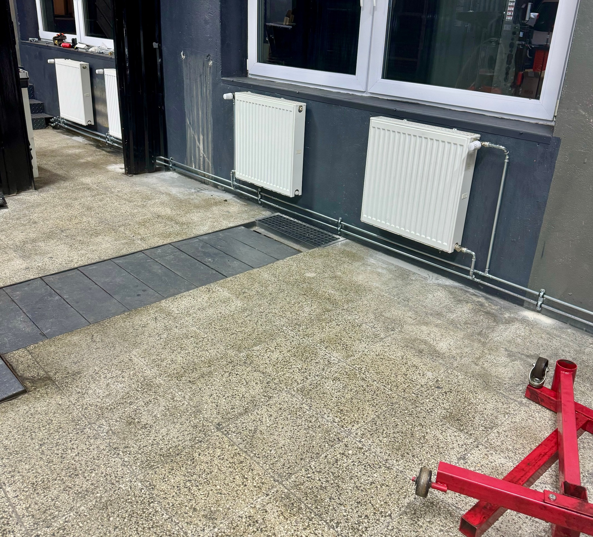 Białe grzejniki panelowe zamontowane na szarej ścianie, z widocznymi rurami i zaworami, na podłodze z terakoty. W tle okno i czarny portal drzwiowy. W rogu czerwony stojak warsztatowy na kółkach.