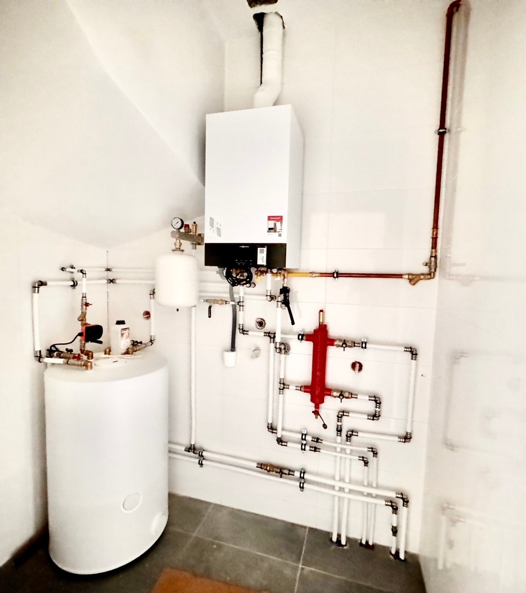 Biały piec centralnego ogrzewania z podłączonymi rurami, zbiornik wody i rozdzielacz w kotłowni z białymi ścianami i szarą podłogą.