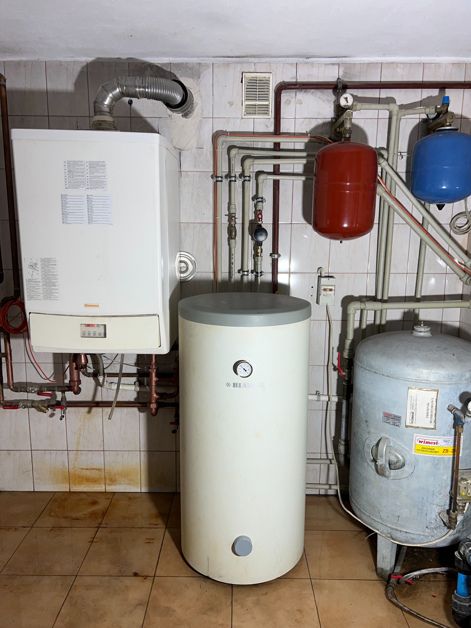 Kotłownia z piecem gazowym, zasobnikiem CWU Biawar, zbiornikami wyrównawczymi i instalacją rurową w pomieszczeniu z kafelkami na ścianach i podłodze.