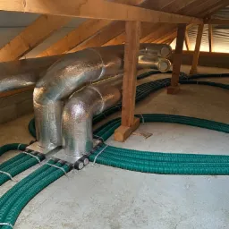 Instalacja rekuperacji z zielonymi rurami w surowym pomieszczeniu pod drewnianą konstrukcją dachu.