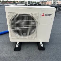 Kompaktowa jednostka zewnętrzna klimatyzacji Mitsubishi Electric, zamontowana na dachu pokrytym papą, widoczne czarne podkładki antywibracyjne i fragment niebieskiego węża.