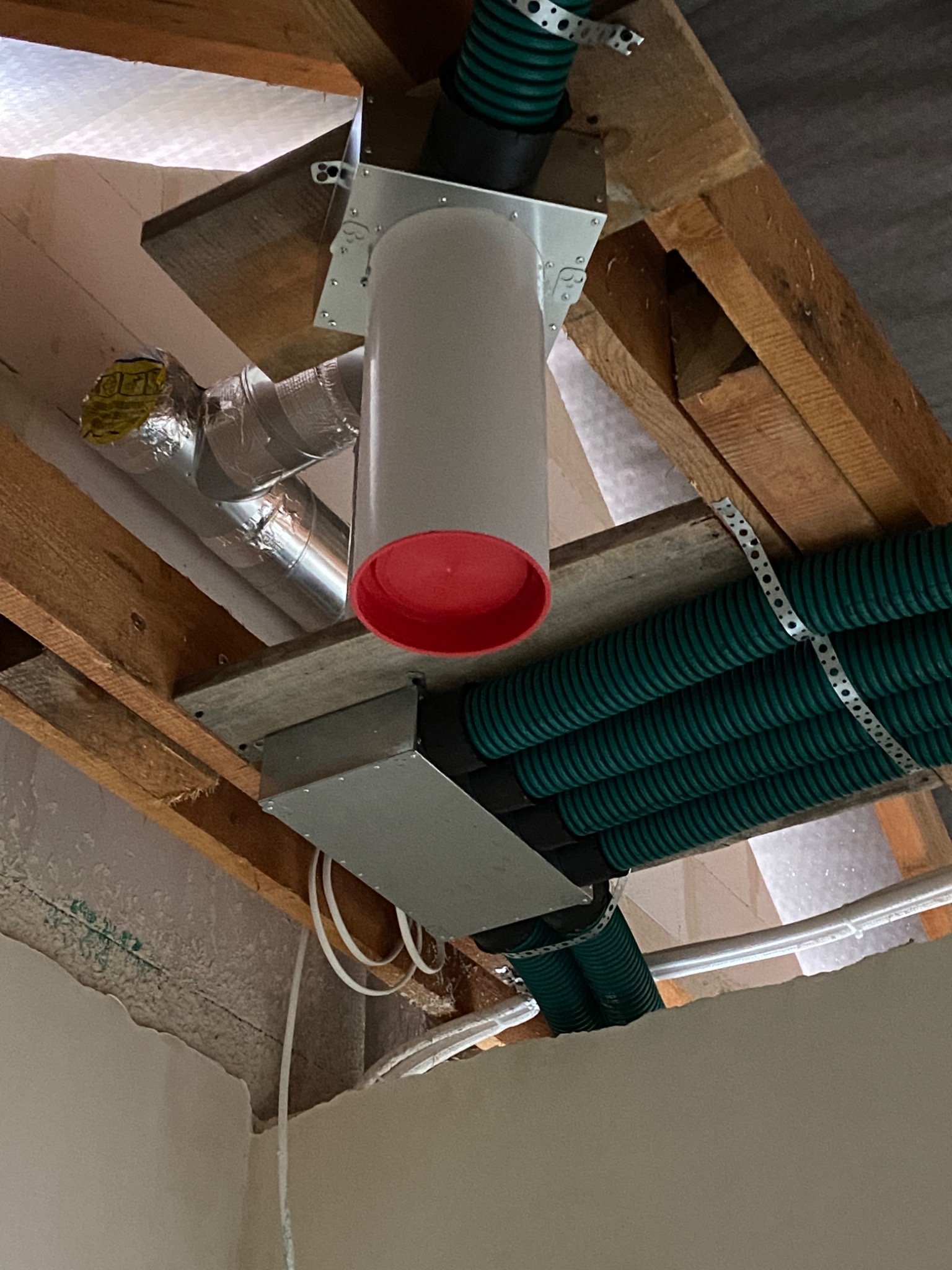 Fragment instalacji wentylacyjnej pod sufitem, widoczne rury wentylacyjne w kolorze zielonym, elementy metalowe mocujące, biały przewód z czerwonym zakończeniem oraz drewniana konstrukcja stropu.