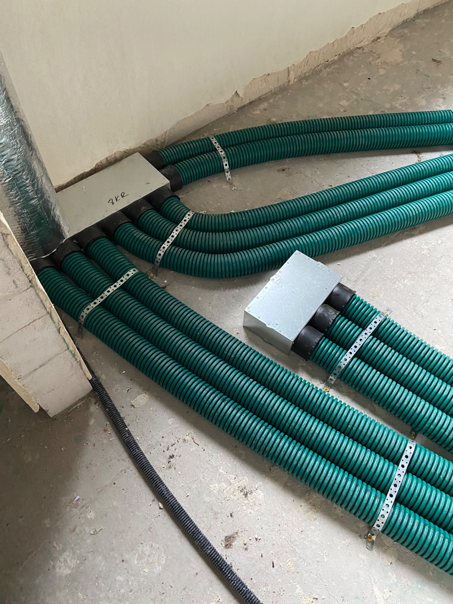 Zielone, karbowane rury wentylacyjne przymocowane metalowymi obejmami do podłoża, wchodzące do metalowych puszek rozdzielczych. Widoczny fragment instalacji z folią aluminiową.