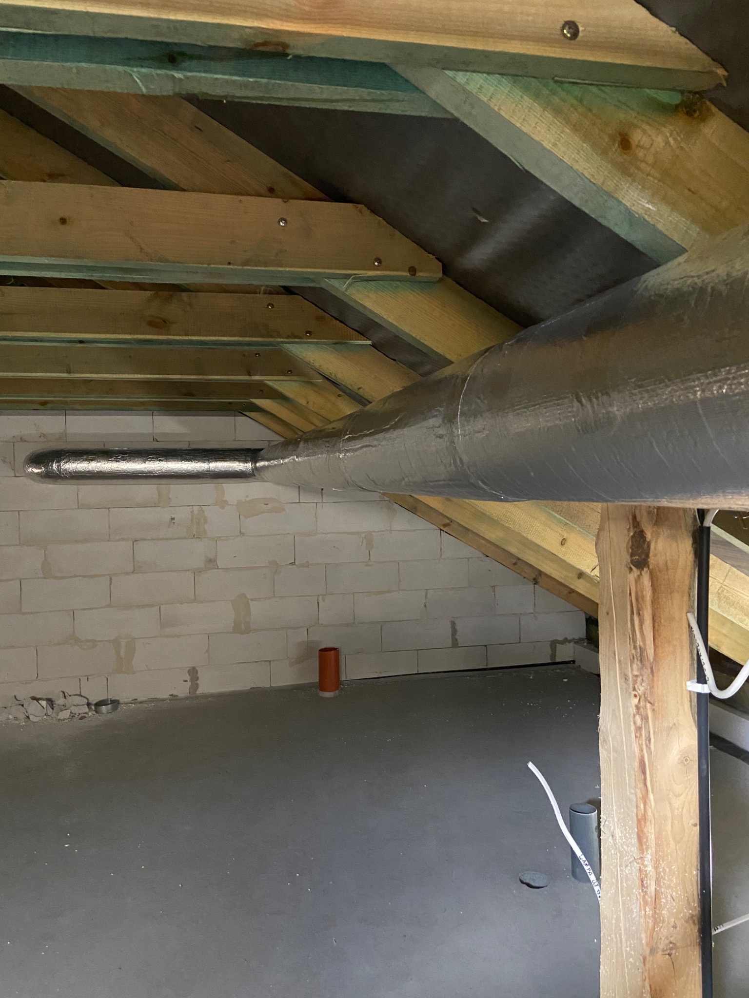 Szary, izolowany przewód wentylacyjny przechodzi przez poddasze z surowymi ścianami z bloków i drewnianą konstrukcją dachu.